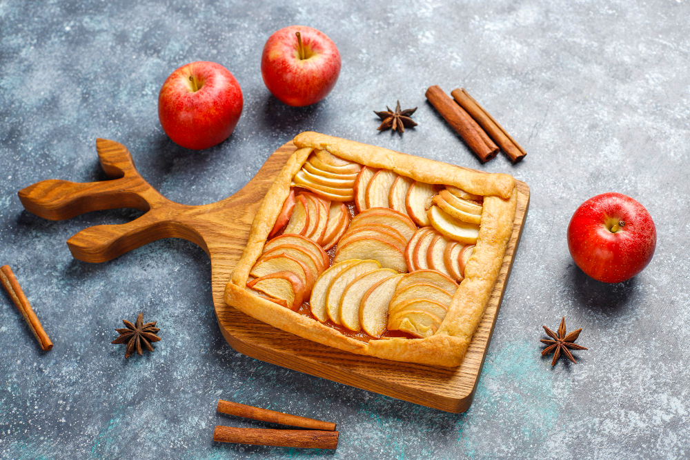 A top-down view of a rustic, square apple tart with a braided crust on a wooden cutting board shaped like a tree. The tart is filled with thinly sliced apples arranged in a circular pattern. Two whole red apples, cinnamon sticks, and star anise pods are scattered around the board on a blue-grey textured surface.