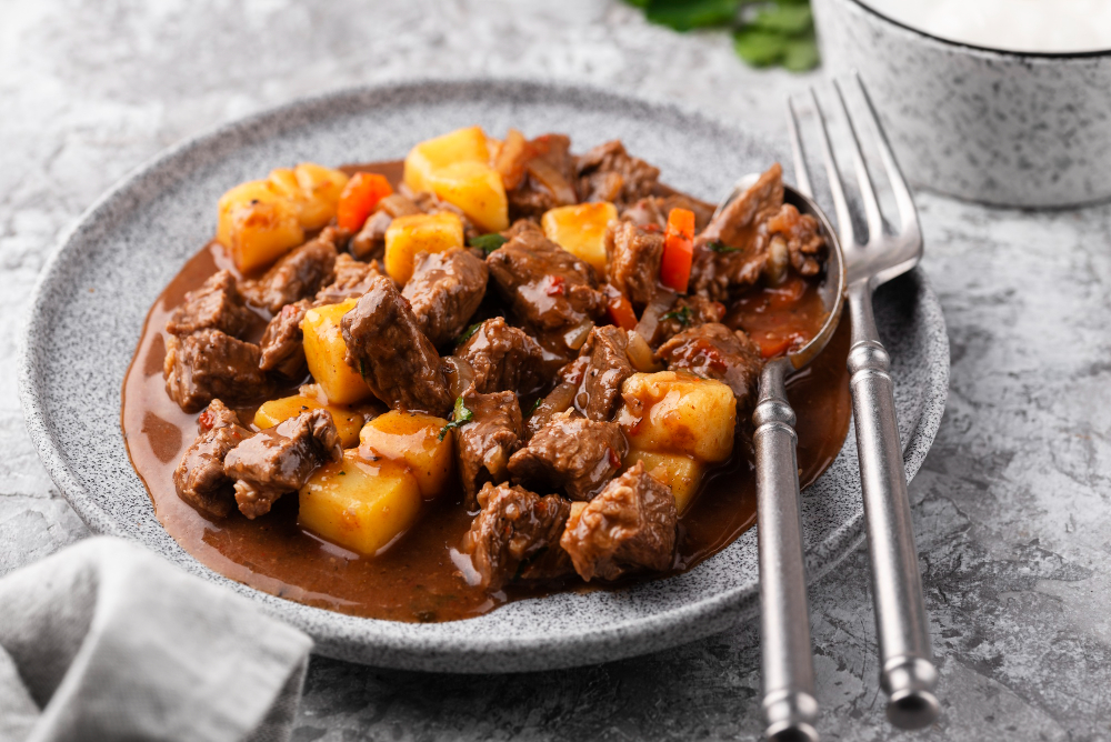 A grey speckled plate holds a serving of savory braised beef and potato stew. The chunky beef and cubed potatoes are in a rich, dark gravy. A fork and a spoon rest on the side of the plate, which is placed on a stone-like surface. A white linen napkin is tucked under the plate on the left.