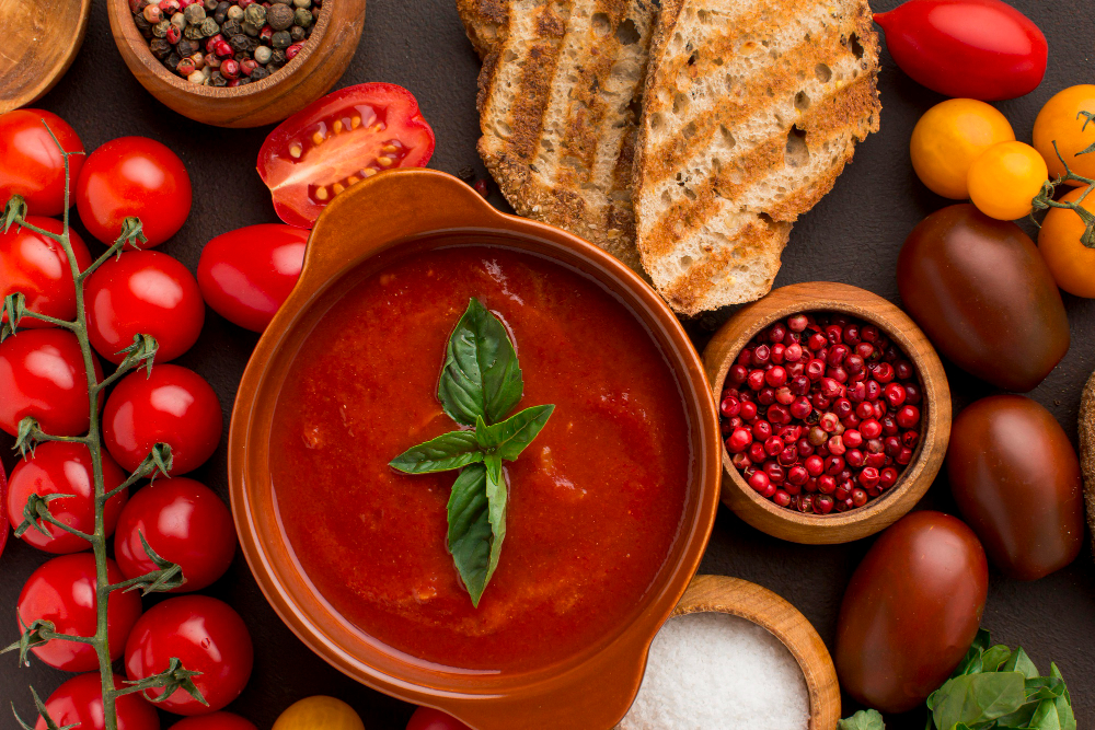 A terracotta bowl of smooth tomato soup, garnished with a single basil leaf, sits in the center of a dark surface. Toasted rustic bread slices are placed above the bowl. The scene is surrounded by various ingredients, including a vine of red cherry tomatoes, an assortment of different colored heirloom tomatoes, a bowl of mixed peppercorns, a bowl of pink peppercorns, and a small wooden bowl of sea salt.